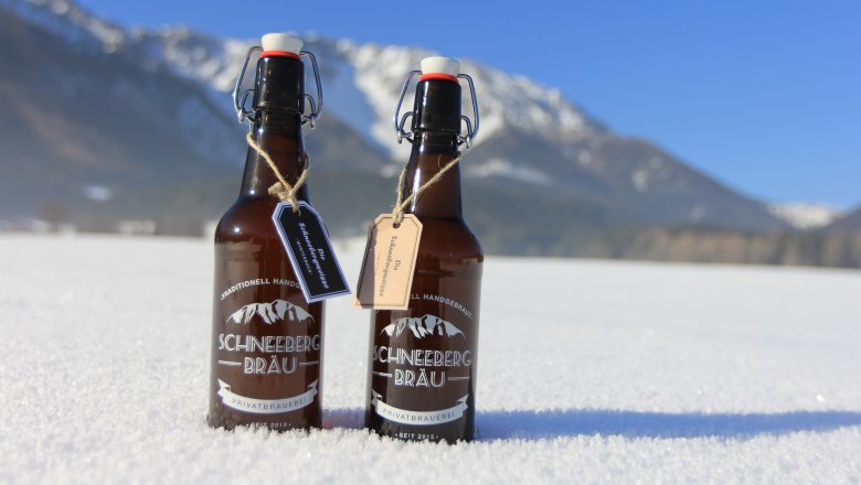 Two bottles of Schneeberg Br&auml;u beer stand in the snow against a mountain backdrop.