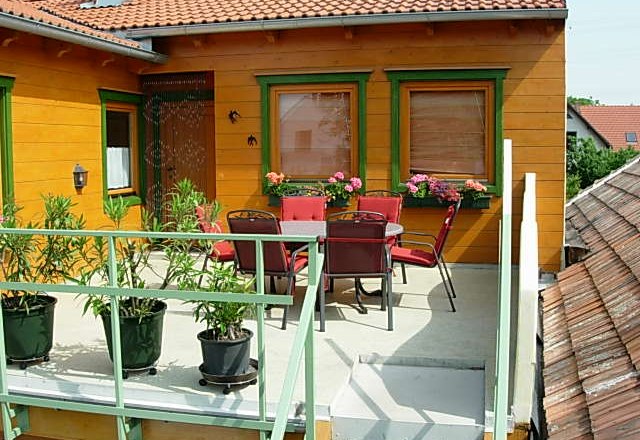 Balcony with table, chairs and plants, surrounded by a house with an orange façade and green window frames.