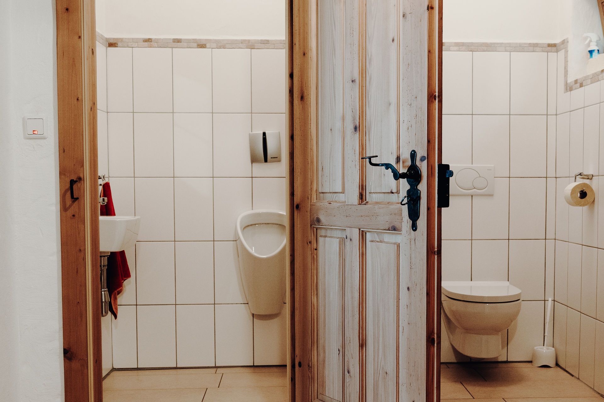 Two toilet rooms with wooden doors, a urinal and washbasin on the left, a toilet on the right.