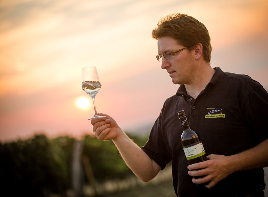 A man holds a wine glass and a wine bottle in the vineyard at sunset.