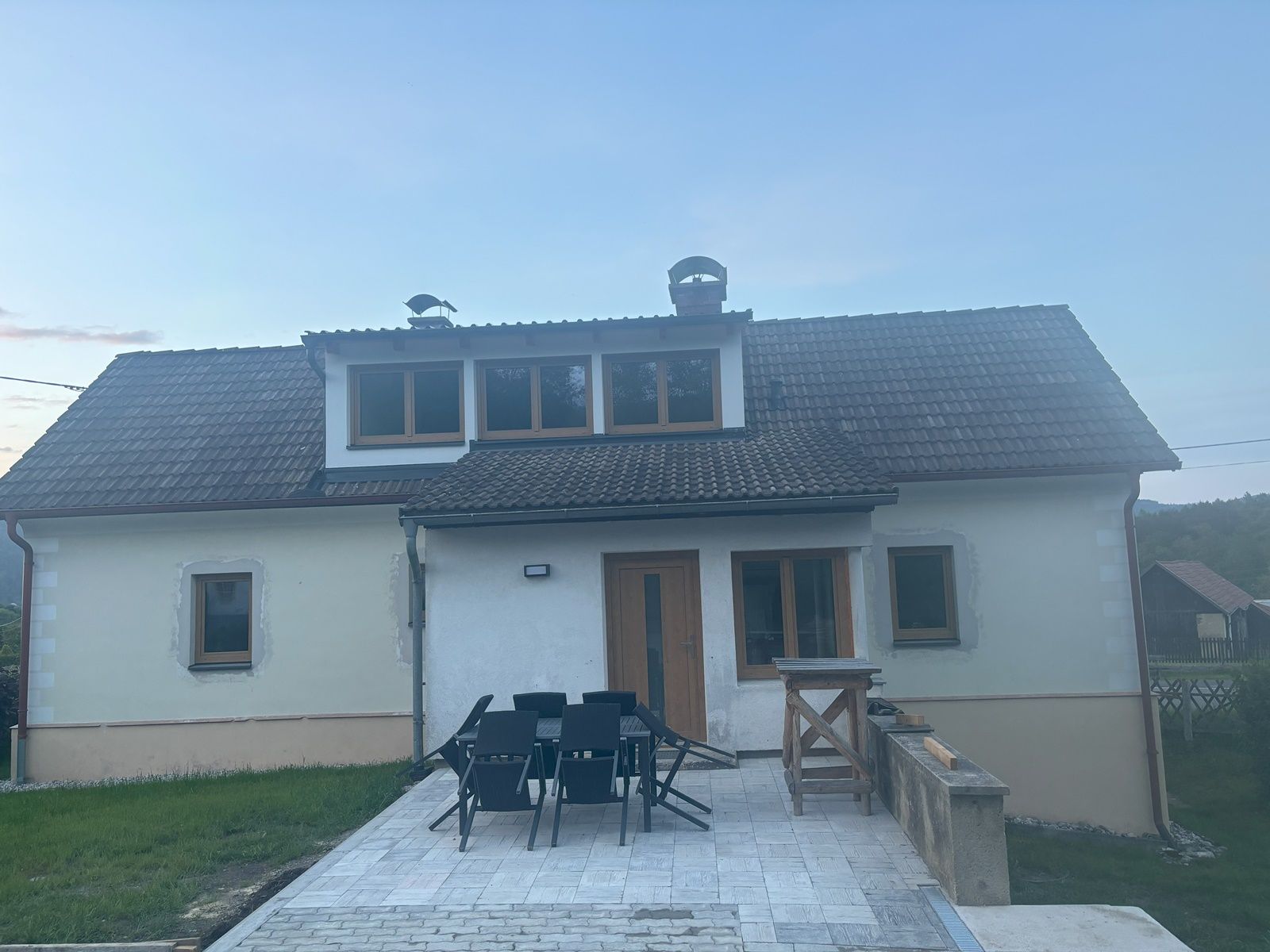 A two-storey cottage with shingled roof and terrace, surrounded by lawn and rural surroundings.