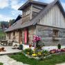 A rustic wooden hut with a flower bed and a dog on the terrace, surrounded by green countryside and red parasols.