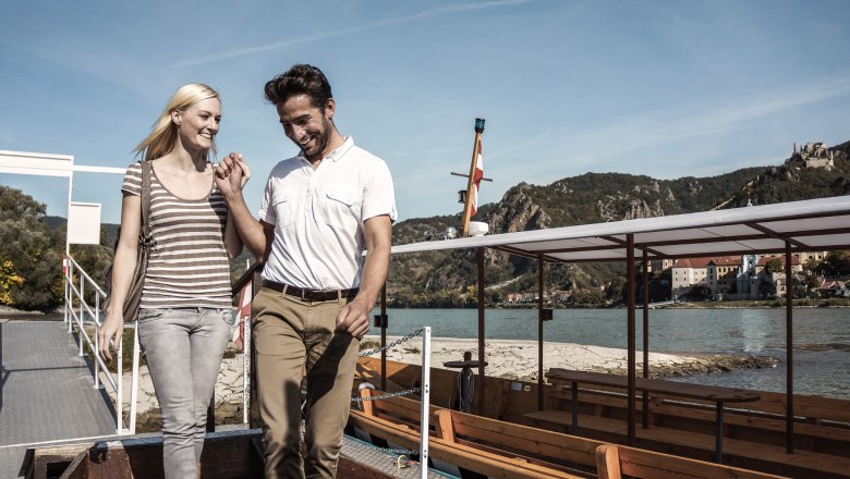 A smiling couple gets out of a boat on a riverbank, mountains and buildings can be seen in the background.