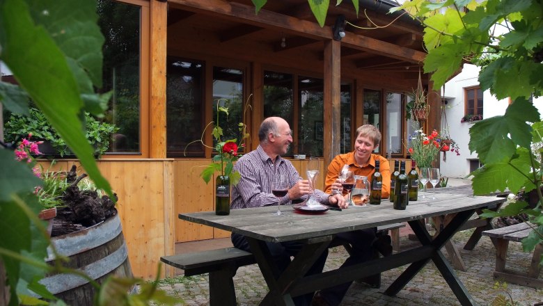 Wine tasting, © www.seymann-film.at Two men sit at a wooden table outside and taste wine. Surrounded by plants and a wooden building in the background.