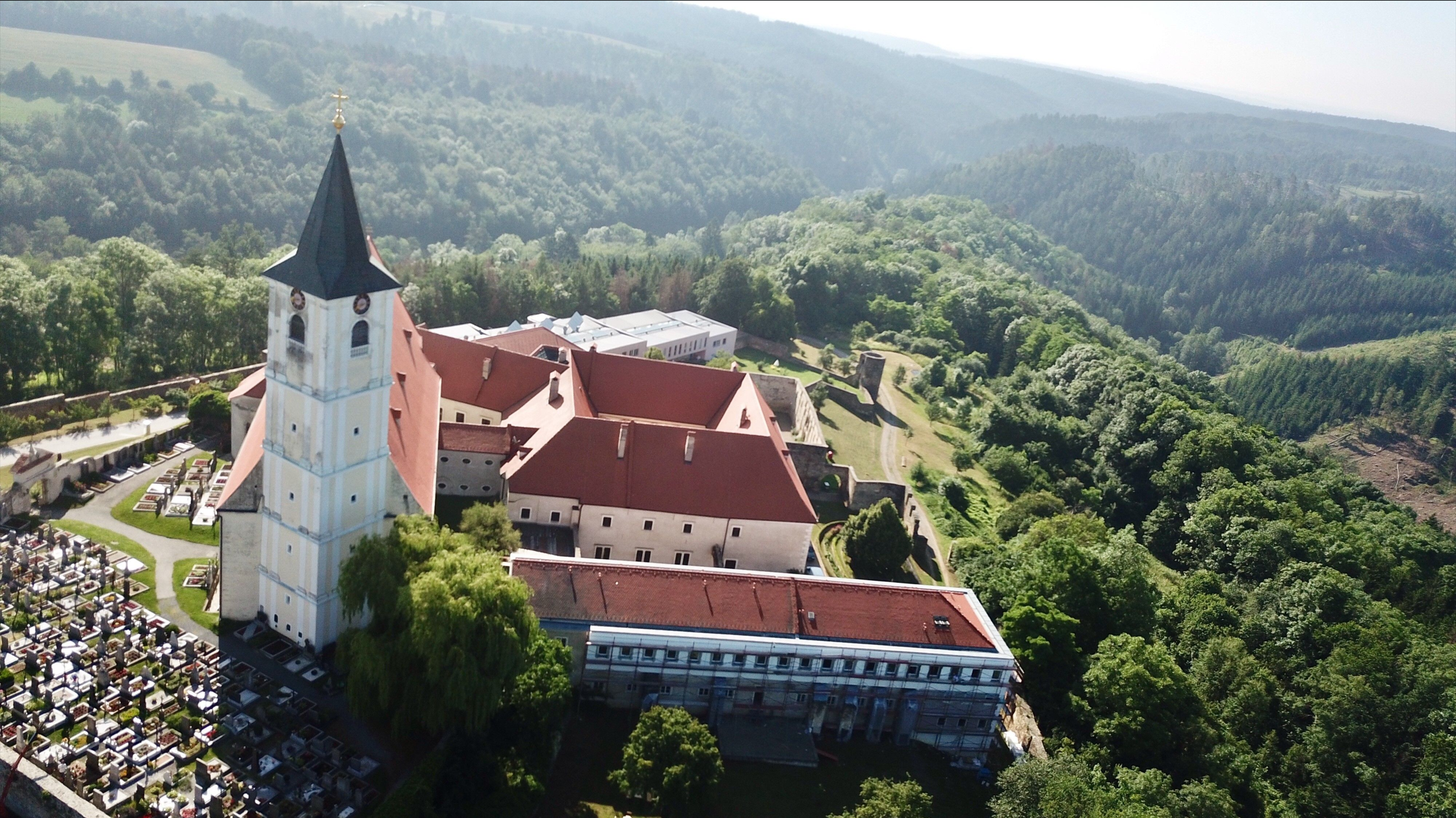 Aerial view of a monastery with a red roof and a cemetery next to it, surrounded by wooded hills.
