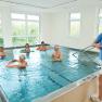 People in an exercise pool with swimming noodles, supervised by a trainer.