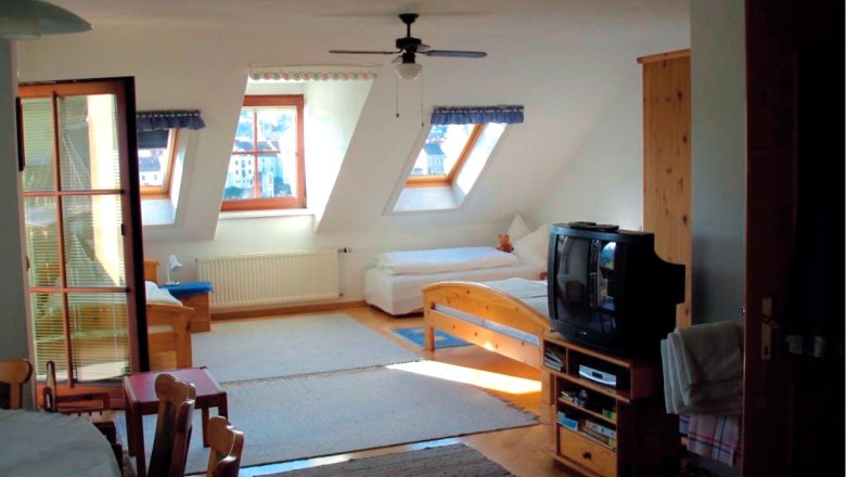 Guesthouse Tüchler, © Gästehaus Tüchler Interior view of a cozy attic room with wooden furniture, two beds, a TV and skylights.