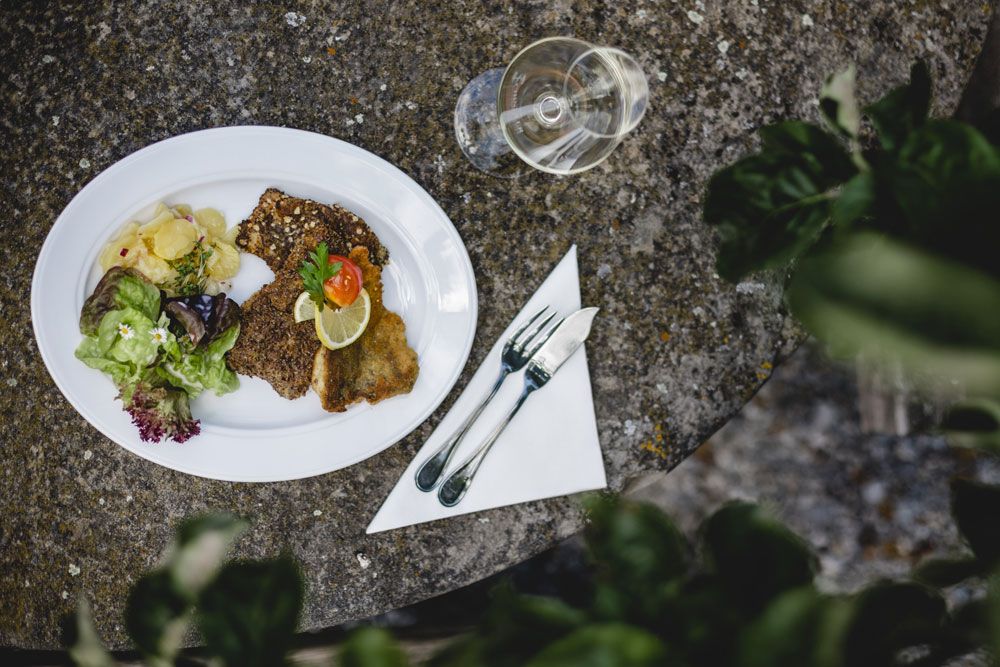 A plate with breaded fish, salad and potatoes on a stone table, cutlery and a wine glass next to it.