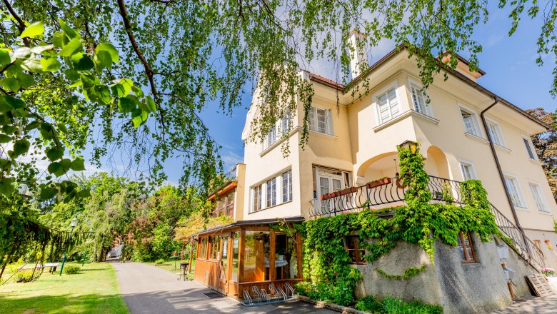 Woltron Manor, © Wiener Alpen / Christian Kremsl A yellow manor house overgrown with ivy, surrounded by trees and a garden path.