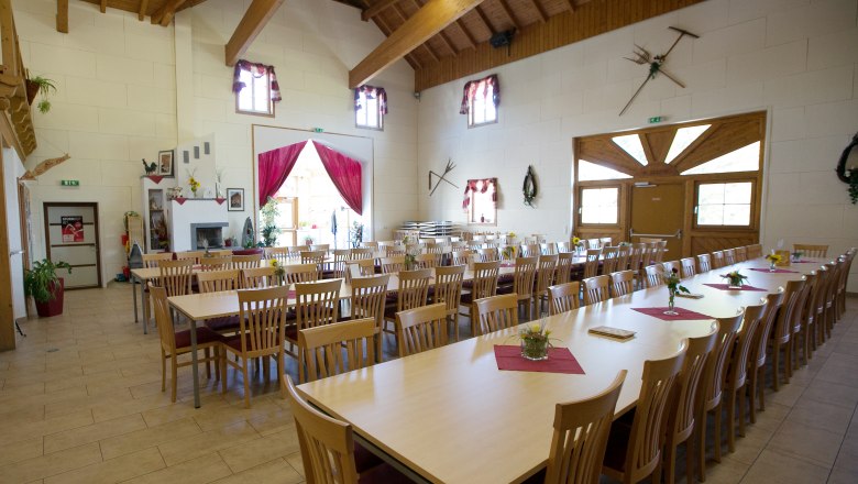A large, bright room with long wooden tables and chairs, decorated with flowers and red table runners, under a wooden roof.