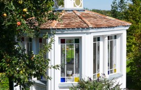 A white pavilion with colorful windows in a garden, surrounded by trees and a fence.