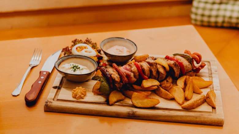 A wooden board with a meat skewer, potato wedges, two sauce bowls and salad on a table.