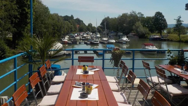 Terrace with wooden tables and chairs, view of a river with boats.