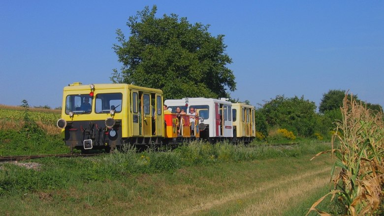 A yellow rail vehicle drives through a rural area with trees and fields.