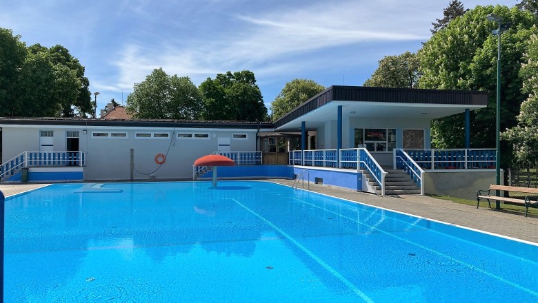 An outdoor pool with clear water, surrounded by trees and a building with blue railings.