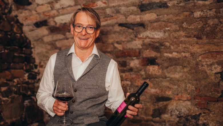 A man with glasses holds a wine glass and a wine bottle in front of a brick wall.