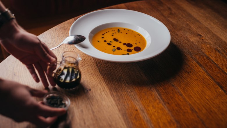 A plate of pumpkin soup with oil on a wooden table, hands with spices next to it.
