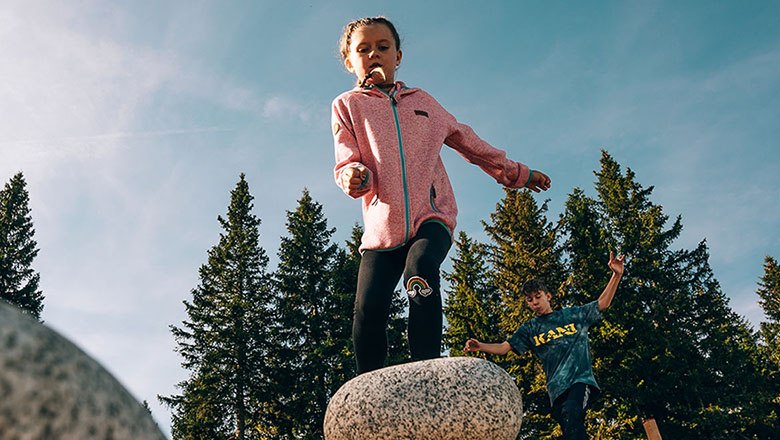 Children play on wobbly stones against a backdrop of trees and blue sky.