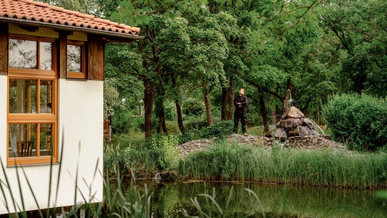 A man stands next to a small fountain in a green garden with a pond and a building with red roof tiles.