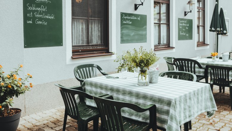 A guest garden with green chairs and checkered tablecloths in front of a house wall with chalkboards.