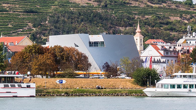 Modern architecture of the Landesgalerie Niederösterreich on the riverbank with surrounding buildings and boats.
