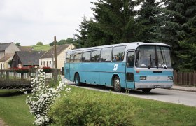 A blue bus drives through a village with flowers and trees in the background.