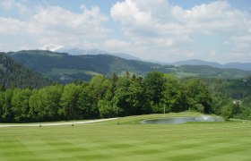 Green golf course landscape with pond and wooded hills in the background.