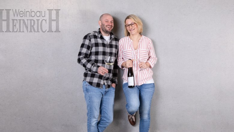 Two people stand smiling in front of a gray wall with wine glasses in their hands. On the left is a man, on the right a woman. At the top left it says 'Weinbau Heinrich'.