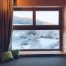 View from a wooden window of a snowy landscape with mountains in the background.