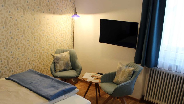 Zimmer mit Bett, Tisch mit zwei Stühlen, Fernseher, Fenster mit blauen Vorhängen und Stehlampe., © Magdalena Donhauser