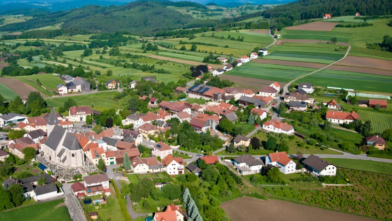 Aerial view of a village with a church, surrounded by green fields and hills.