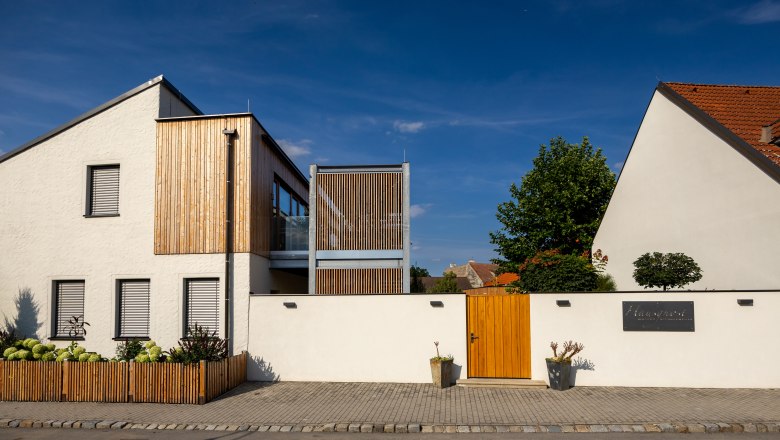 Modern building with a white façade and wooden elements, surrounded by a wall with a wooden gate.