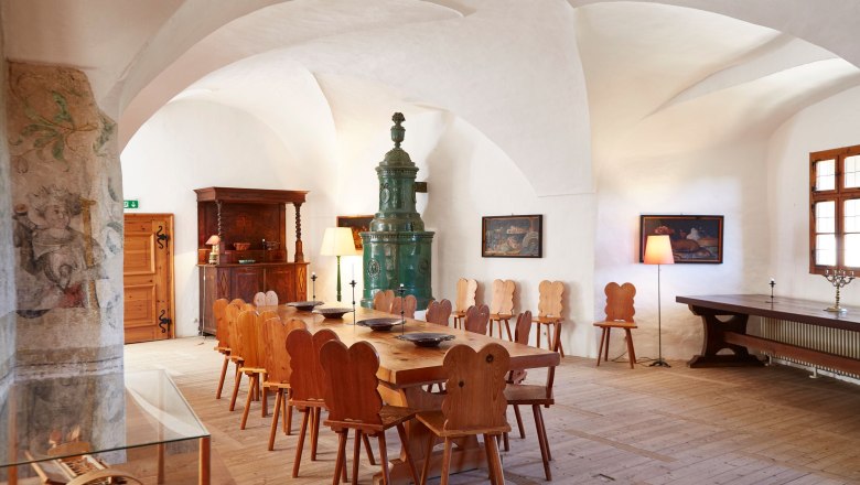 Interior view of a historic room with wooden table and chairs, a green tiled stove and wall paintings.