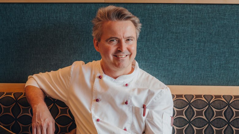 A smiling man in a white chef's jacket sits on an upholstered bench in front of a patterned wall.