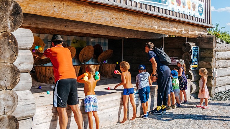 Children and adults throw colorful balls in an outdoor wooden play stand.