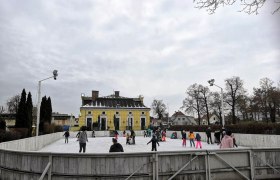 People skating on an open-air rink in front of a yellow building.