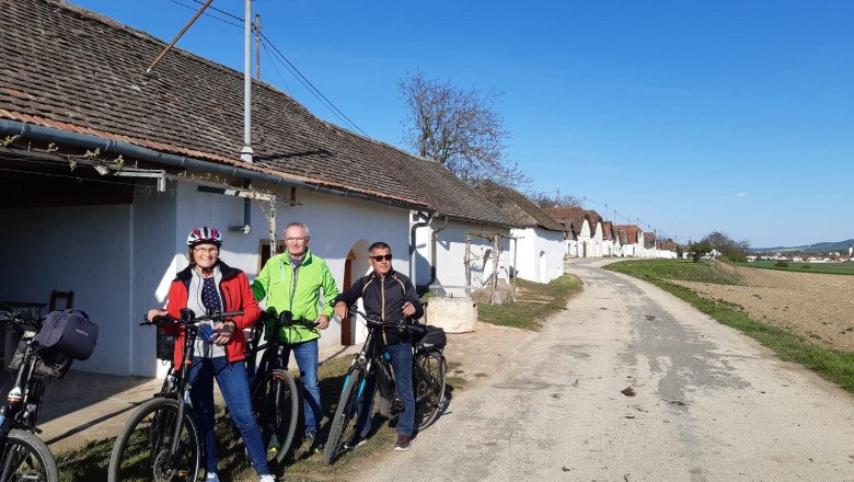 Three people with bicycles in front of a row of wine cellars in a rural setting.