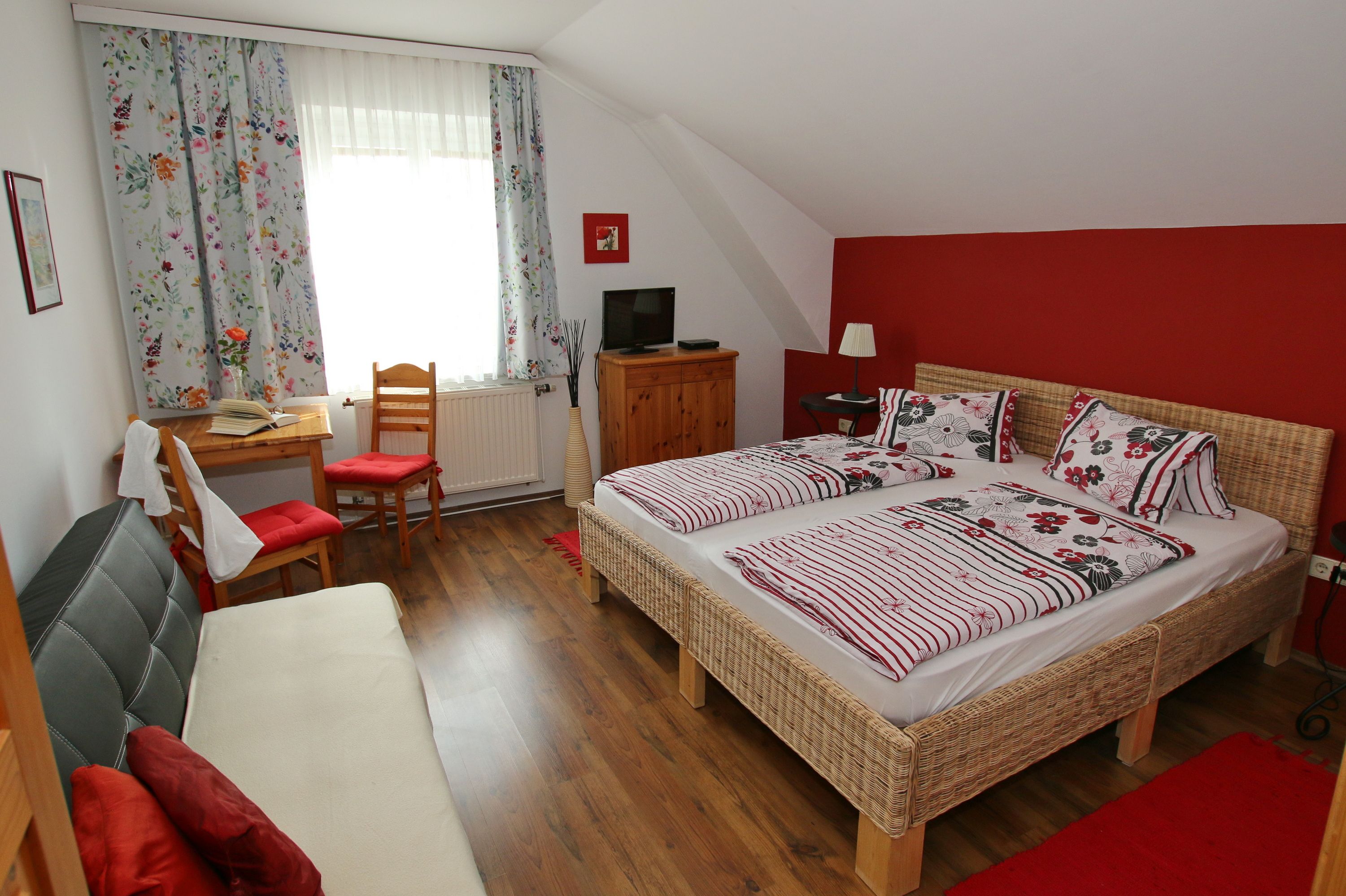 Cozy guest room with double bed, sofa, table and chairs. Red accent wall and floral curtains.