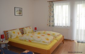 A cozy double room with a wooden bed, colorful bedding and two red bedside lamps. A window with curtains is in the background.