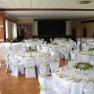 Elegant wedding hall with round tables, white tablecloths and chair covers, decorated with green plants and folded napkins.