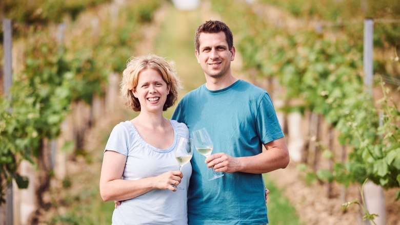 A smiling couple stands in a vineyard holding glasses of wine.