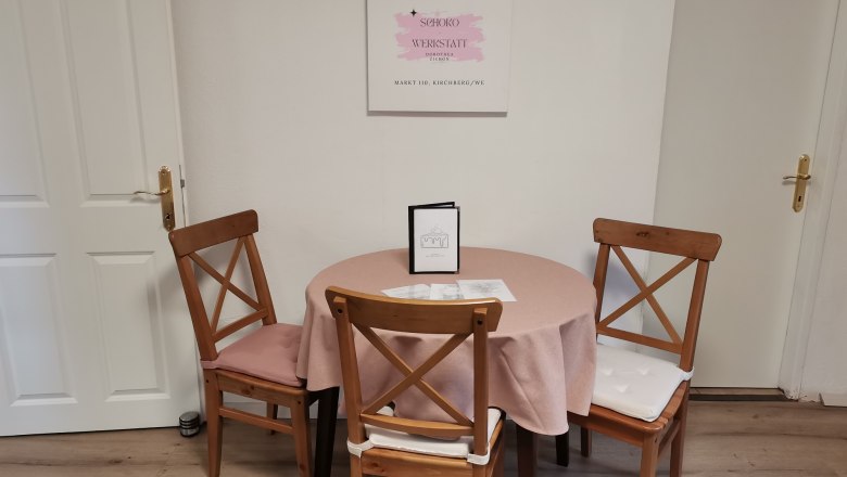 A table with a pink tablecloth and three wooden chairs in a café, with a sign on the wall.