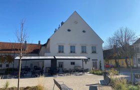 A garden with empty tables and chairs in front of a white building with a pointed roof on a sunny day.