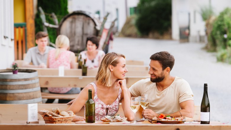 A couple is sitting at an outdoor table, enjoying wine and food. Other guests and a wine barrel can be seen in the background.
