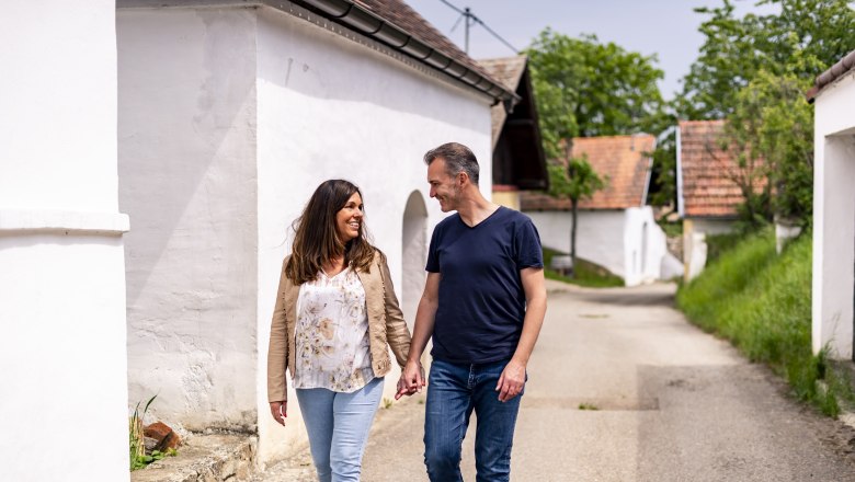 A couple walks hand in hand through the &Ouml;hlbergkellergasse in Pillersdorf, surrounded by traditional wine cellars and green countryside.