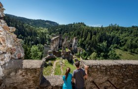 Two people look at the Kollmitz ruins from a wall, surrounded by a dense forest and blue sky.