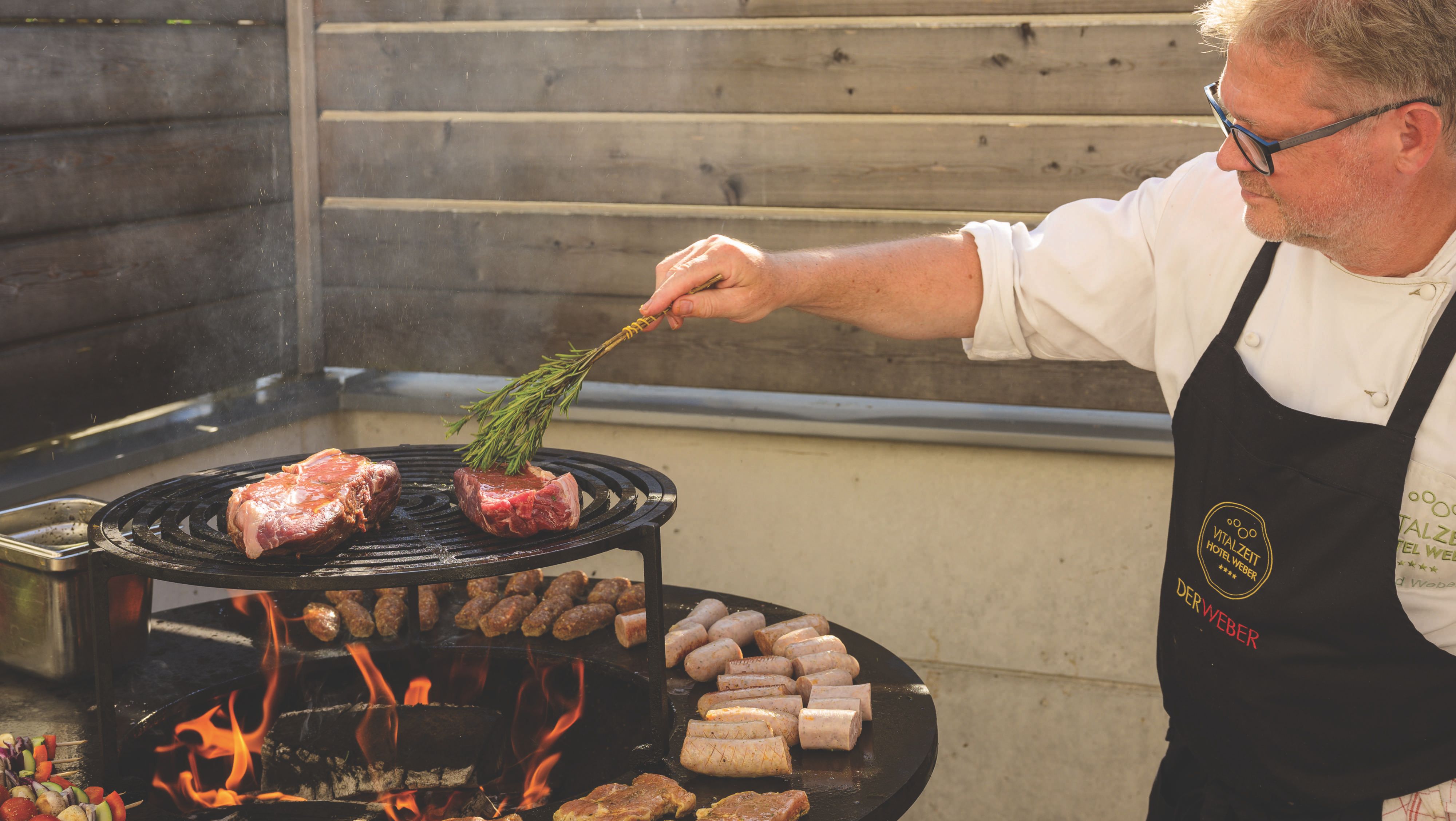 The owner of the restaurant is grilling meat and sausages on a round grill over an open fire.