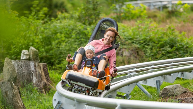 A woman and a child ride on a summer toboggan run through a green landscape.