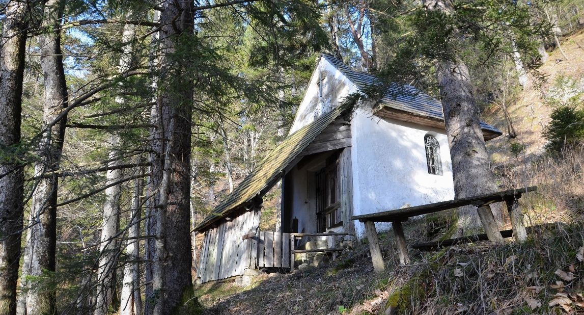 A small white chapel in the forest, surrounded by trees and a wooden table in the foreground.